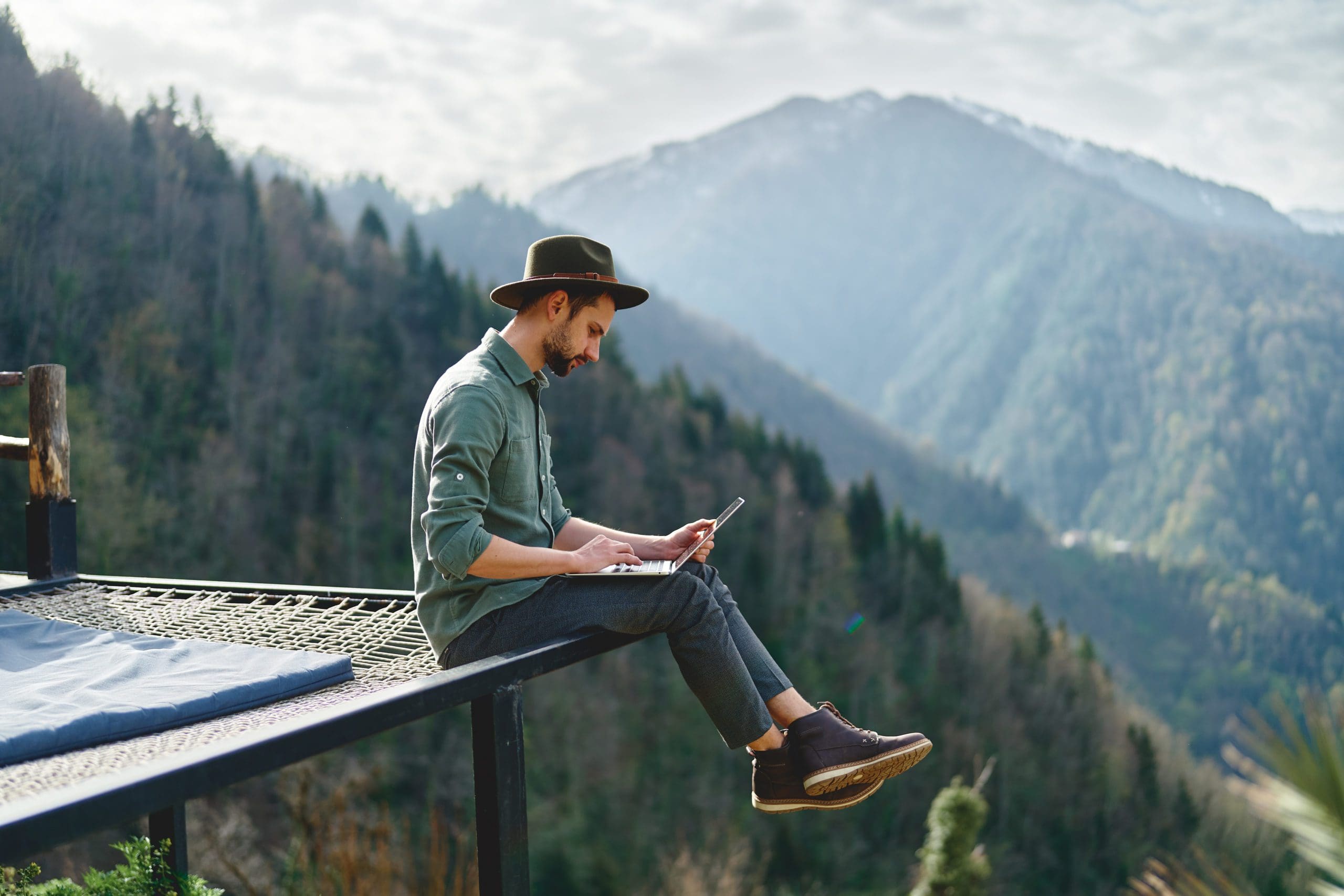 Young man freelancer traveler wearing hat anywhere working online using laptop and enjoying mountains view hybrid mail